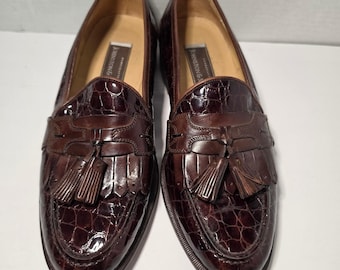 May include: A pair of dark brown loafers featuring a glossy, embossed crocodile-patterned leather finish. Each shoe is adorned with a decorative fringe kiltie and a matching leather tassel detail on the vamp. The interior label reads Johnston & Murphy.