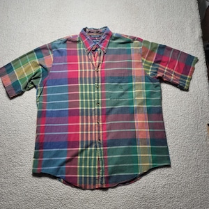 May include: A short-sleeved button-down shirt featuring a multi-coloured plaid pattern in shades of red, green, blue, and yellow. The shirt includes a pointed collar, a front button placket, and a single chest pocket with an embroidered logo.