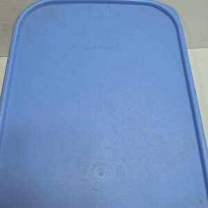 May include: A rectangular light blue plastic storage container lid with rounded corners. The word Tupperware is embossed in the center near the top edge, and a circular manufacturing mark is visible toward the bottom center of the surface.
