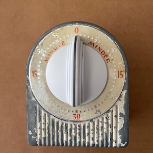 May include: A vintage kitchen timer with a grey metal base featuring chipped paint and a white central dial. The face is cream-colored with red text reading MINUTE MINDER and black numbers from 0 to 55. The dial has a vertical ribbed texture.