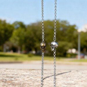 Puede incluir: Dos collares de cadena plateados, cada uno con un colgante de calavera. Una calavera es dorada, la otra es plateada. Las cadenas cuelgan sobre un fondo borroso de &aacute;rboles verdes y cielo azul. Los collares se exhiben al aire libre.