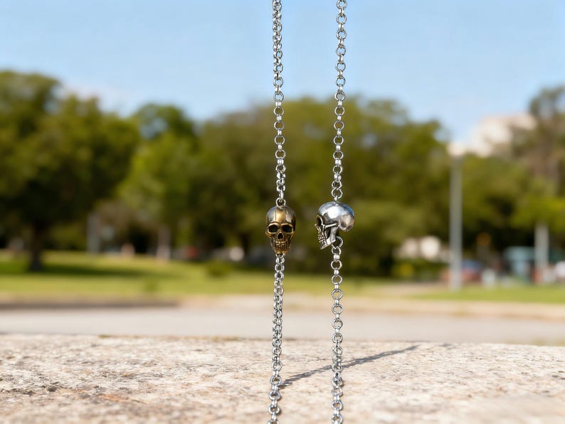 Puede incluir: Dos collares de cadena plateados, cada uno con un colgante de calavera. Una calavera es dorada, la otra es plateada. Las cadenas cuelgan sobre un fondo borroso de &aacute;rboles verdes y cielo azul. Los collares se exhiben al aire libre.