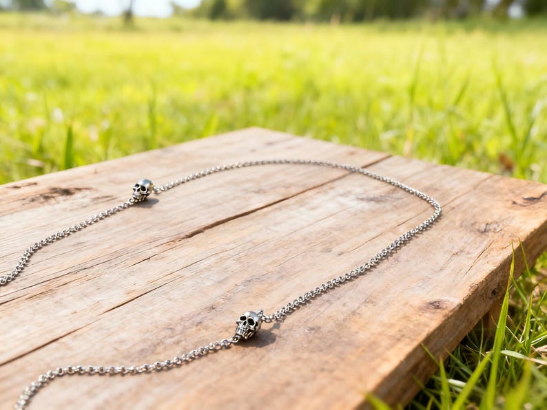 Puede incluir: Un collar plateado con colgantes de calavera. La cadena est&aacute; hecha de peque&ntilde;os eslabones redondos. Las calaveras tienen cuencas de los ojos y una cavidad nasal detalladas. El collar se muestra sobre una superficie de madera envejecida.