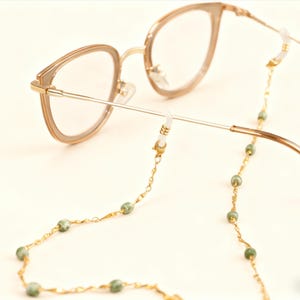 May include: Gold-toned eyeglasses with a matching chain. The chain features small, green beads and connects to the glasses' temples. The glasses have a light brown frame and clear lenses. The chain is a stylish accessory.