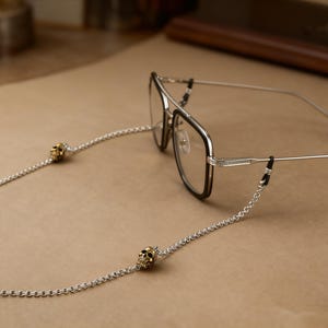 Puede incluir: Gafas con una cadena plateada con detalles de calaveras doradas. Las gafas tienen una montura negra y plateada. La cadena est&aacute; sujeta a las patillas de las gafas. El fondo es una superficie marr&oacute;n claro.