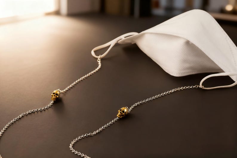 Puede incluir: Una mascarilla blanca con una cadena plateada con detalles de calaveras doradas. La mascarilla est&aacute; hecha de un material suave y transpirable. La cadena est&aacute; dise&ntilde;ada para sujetar la mascarilla alrededor del cuello.