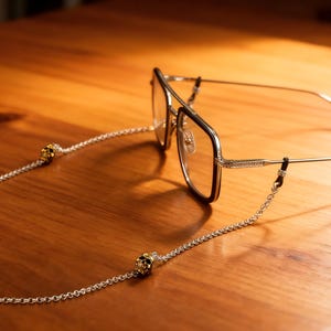 Puede incluir: Gafas con una cadena plateada y adornos de calavera sobre una superficie de madera. Las gafas tienen una montura dorada con un borde oscuro. La cadena est&aacute; sujeta a las patillas de las gafas.
