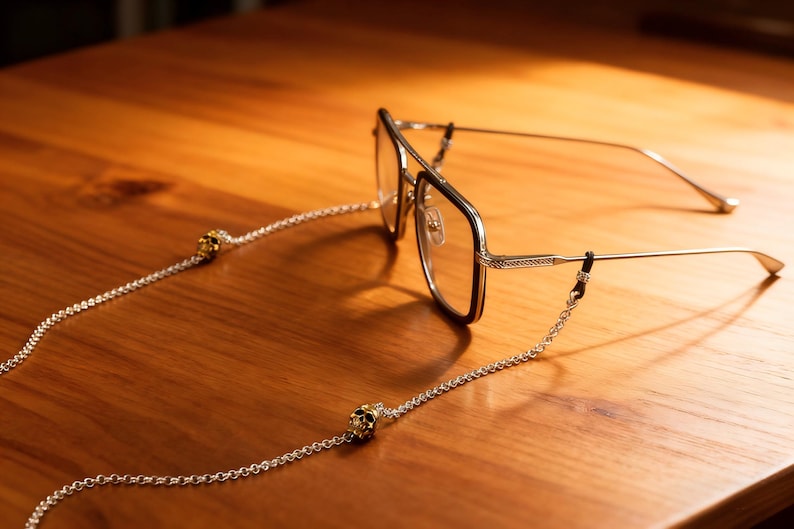 Puede incluir: Gafas con una cadena plateada y adornos de calavera sobre una superficie de madera. Las gafas tienen una montura dorada con un borde oscuro. La cadena est&aacute; sujeta a las patillas de las gafas.