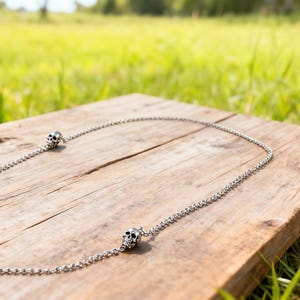 Puede incluir: Un collar plateado con colgantes de calavera. La cadena est&aacute; hecha de peque&ntilde;os eslabones redondos. Las calaveras tienen cuencas de los ojos y una cavidad nasal detalladas. El collar se exhibe sobre una superficie de madera envejecida.