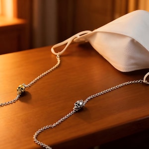 Puede incluir: Una mascarilla blanca con una cadena plateada con abalorios de calaveras. La cadena est&aacute; unida a las presillas de la mascarilla. La mascarilla y la cadena descansan sobre una superficie de madera.