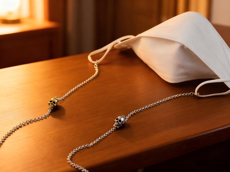 Puede incluir: Una mascarilla blanca con una cadena plateada con abalorios de calaveras. La cadena est&aacute; unida a las presillas de la mascarilla. La mascarilla y la cadena descansan sobre una superficie de madera.