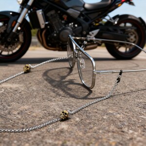Puede incluir: Gafas con montura plateada con una cadena con detalles de calaveras doradas. Las gafas descansan sobre una superficie texturizada, con una motocicleta negra al fondo. La cadena est&aacute; sujeta a las patillas de las gafas.
