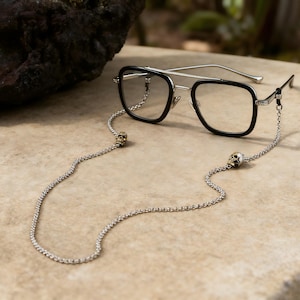 Puede incluir: Gafas negras y plateadas con una cadena con detalles de calaveras. Las gafas tienen montura negra y lentes transparentes. La cadena es plateada y tiene un dije de calavera en cada extremo.