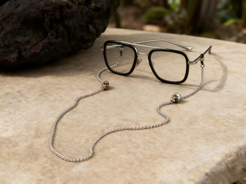 Puede incluir: Gafas negras y plateadas con una cadena con detalles de calaveras. Las gafas tienen montura negra y lentes transparentes. La cadena es plateada y tiene un dije de calavera en cada extremo.