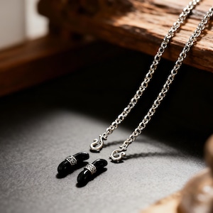 Puede incluir: Primer plano de una cadena plateada con dos soportes para gafas negros de forma ovalada. Cada soporte tiene una banda plateada con un patr&oacute;n decorativo. La cadena est&aacute; hecha de eslabones peque&ntilde;os.