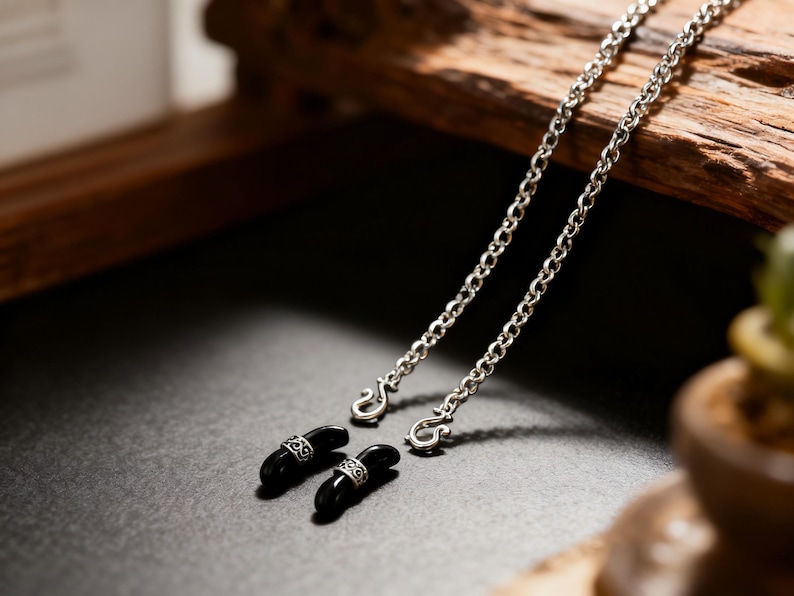 Puede incluir: Primer plano de una cadena plateada con dos soportes para gafas negros de forma ovalada. Cada soporte tiene una banda plateada con un patr&oacute;n decorativo. La cadena est&aacute; hecha de eslabones peque&ntilde;os.