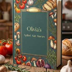 May include: An emerald green cookbook titled "Olivia's Spilled Salt Stories" with illustrations of tomatoes, mushrooms, bread, and herbs. The book has a gold frame and is on a wooden surface.