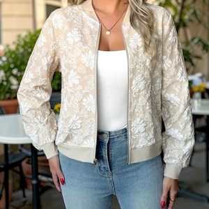 Puede incluir: Chaqueta bomber beige con bordado floral blanco. La chaqueta tiene cremallera, puños y dobladillo acanalados. La modelo lleva una camiseta sin mangas blanca y vaqueros azules.
