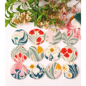 May include: Twelve circular punch needle coasters featuring various floral patterns in shades of red, yellow, pink, white, and green. Each coaster displays a unique botanical design with textured yarn loops arranged in a grid on a white background.