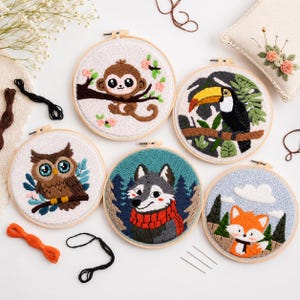 May include: A collection of six embroidered hoops featuring various woodland creatures. The designs include a monkey, toucan, owl, wolf, and fox, each set against a nature-themed backdrop. The embroidery uses a variety of colors and textures.
