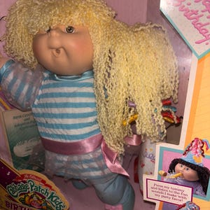 Pu&ograve; includere: Bambola Cabbage Patch Kids Birthday Kids nella sua scatola originale. La bambola ha capelli biondi ricci e indossa un completo a righe blu e bianche con una fascia rosa. La scatola presenta il testo "Happy Birthday" e "Press my tummy and listen to the fun sounds I make with my party favor!"