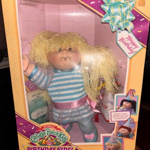 Pu&ograve; includere: Bambola Cabbage Patch Kids Birthday Kids nella sua scatola originale. La bambola ha capelli biondi e ricci e indossa un top a righe blu e bianco, un nastro rosa e pantaloni blu. La scatola ha un'etichetta Happy Birthday e illustrazioni di altre bambole.