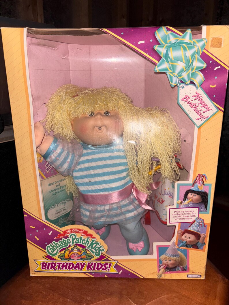 Pu&ograve; includere: Bambola Cabbage Patch Kids Birthday Kids nella sua scatola originale. La bambola ha capelli biondi e ricci e indossa un top a righe blu e bianco, un nastro rosa e pantaloni blu. La scatola ha un'etichetta Happy Birthday e illustrazioni di altre bambole.
