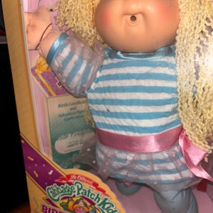 Pu&ograve; includere: Bambola Cabbage Patch Kids nella sua scatola originale. La bambola ha capelli biondi ricci e indossa una maglietta a righe blu e bianche, un nastro rosa e una gonna blu con fiocchi rosa. La scatola dice "Birthday Kids!"