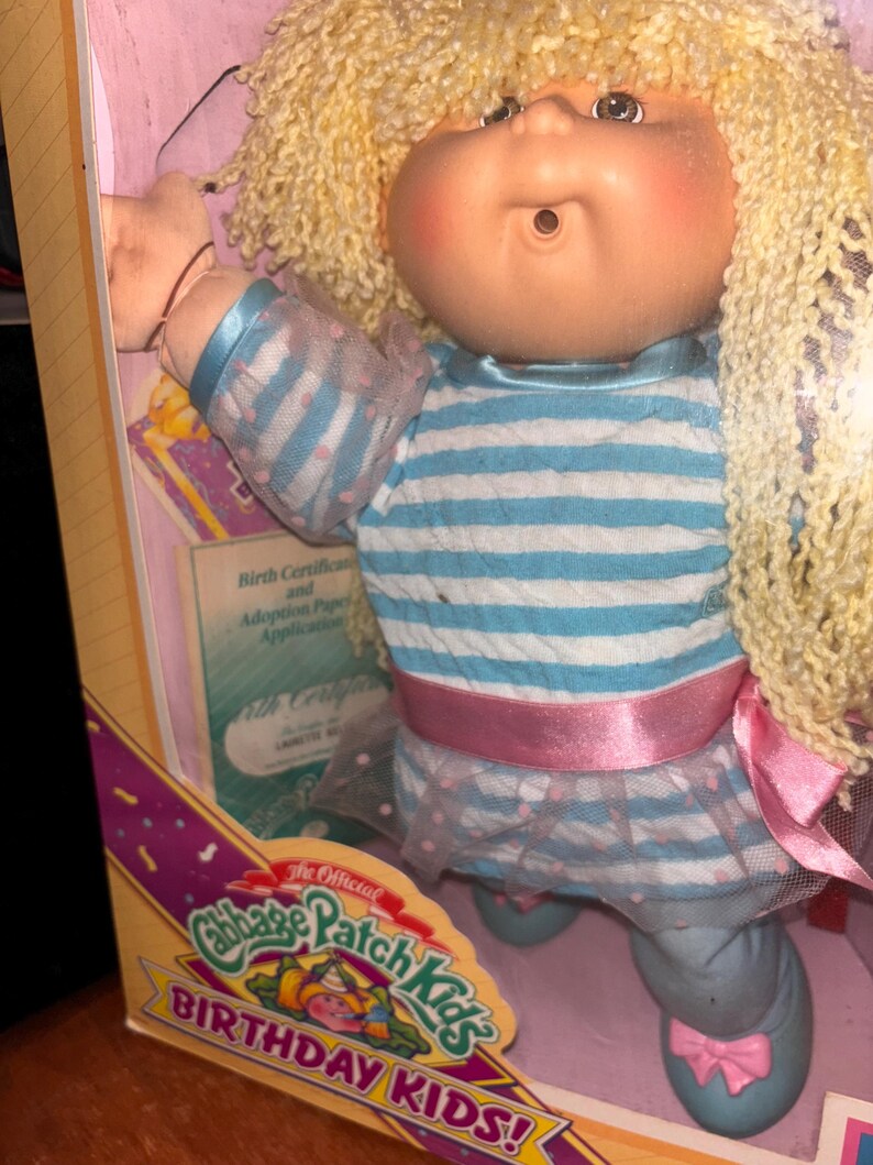 Pu&ograve; includere: Bambola Cabbage Patch Kids nella sua scatola originale. La bambola ha capelli biondi ricci e indossa una maglietta a righe blu e bianche, un nastro rosa e una gonna blu con fiocchi rosa. La scatola dice "Birthday Kids!"