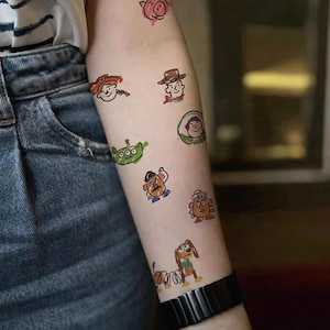 May include: A series of colourful temporary tattoos depicting characters from a well-known animated film. The designs include a pig, a cowboy, a spaceman, a green alien, and other recognisable figures. The tattoos are applied to the arm.