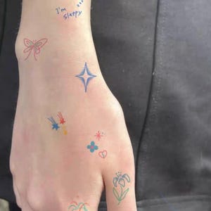 Könnte beinhalten: Eine Hand mit bunten temporären Tattoos. Designs umfassen einen blauen Stern, eine rote Schleife, Sternschnuppen, Blumen, ein Herz und eine Lilie. Die Worte "I'm sleepy" sind mit blauer Tinte in der Nähe des Handgelenks geschrieben. Die Tattoos sind in verschiedenen Farben.