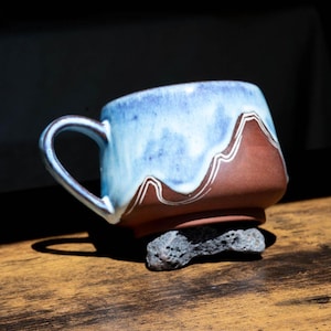 May include: A ceramic mug featuring a matte terracotta brown base and a glossy, drippy light blue glaze on the upper portion. The design includes a white line accent that mimics a mountain range silhouette, with a matching handle and a sturdy base.