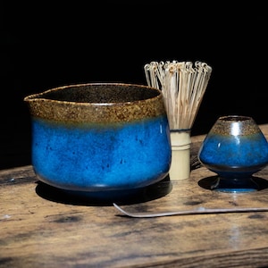Puede incluir: Un cuenco de cerámica para matcha con un esmalte azul vibrante y un borde marrón texturizado, acompañado de un batidor de bambú, un soporte para batidor a juego en azul y marrón, y una cuchara de té de bambú fina y curva sobre madera.