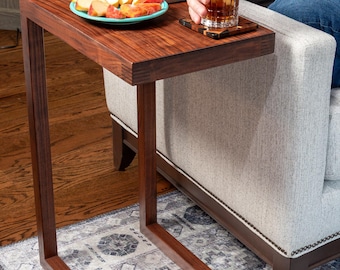 May include: A C-shaped side table constructed from dark brown wood with a visible grain pattern. The table features a flat rectangular top surface supported by a single vertical leg that extends into a horizontal base, forming a continuous frame.