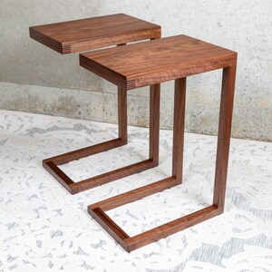May include: Two C-shaped side tables crafted from dark brown wood with a visible grain. Each table features a rectangular top and a single vertical support leg that extends into a flat base, showcasing detailed finger joint accents at the corners.