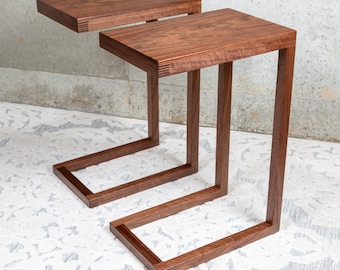 May include: Two C-shaped side tables crafted from dark brown wood with a visible grain. Each table features a rectangular top and a single vertical support leg that extends into a flat base, showcasing detailed finger joint accents at the corners.