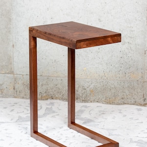 May include: A small, rectangular wooden side table with a warm brown finish. The table has a simple, modern design with a rectangular top and a C-shaped base. The table is approximately 24 inches long, 12 inches wide, and 24 inches high.