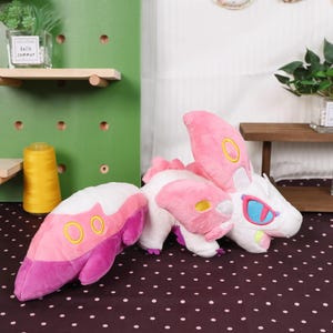 May include: A plush toy dragon in white, pink, and purple. The dragon has large pink wings with yellow accents, blue eyes, and a long tail. The dragon is lying on a brown surface with white polka dots. The background includes shelves and decorative items.