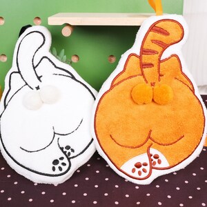 May include: Two cat-shaped plushies, one white and one orange, are displayed. The plushies are embroidered with black outlines and feature the cat's rear end, tail, and paws. Each has a loop for hanging. The orange cat has a striped tail.