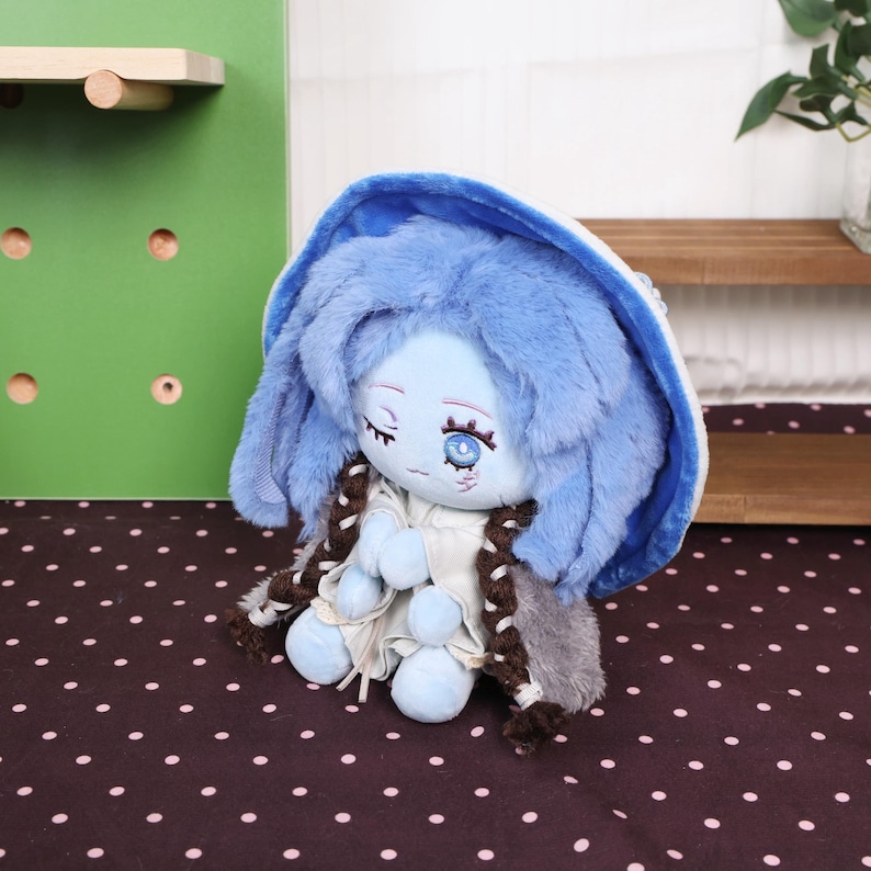 May include: A plush doll with blue hair and a matching hat. The doll has a light blue face with closed eyes and a detailed eye design. It wears a white dress and a gray and brown shawl. The doll is sitting on a brown surface with white polka dots.