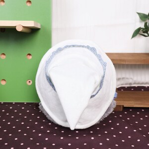May include: A white witch hat with a pointed crown and a wide brim. The brim is trimmed with a delicate blue lace. The hat is set against a backdrop of a green wall and a brown surface with white polka dots.