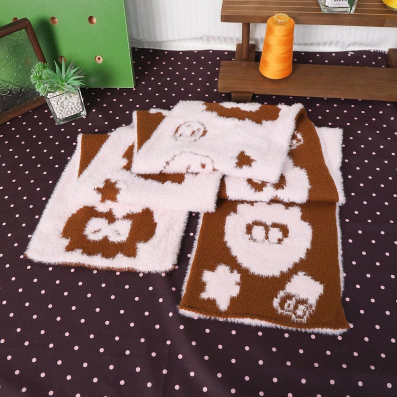 May include: A brown and white scarf with cartoon animal designs. The scarf is made of a soft, fuzzy material and is folded on a polka dot surface. The scarf features a repeating pattern of animal faces and shapes.