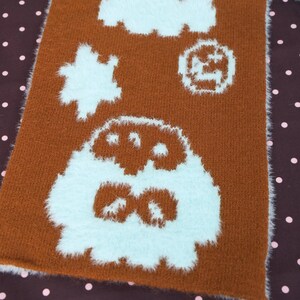 May include: Brown rectangular scarf with white fluffy cartoon-like designs. The scarf has a brown background with white fluffy shapes, including a star, a circle, and a large character at the bottom. The edges of the scarf are also white and fluffy.