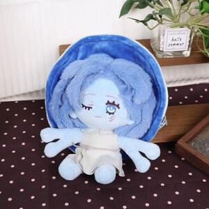 May include: A plush doll with blue skin, hair, and a matching hat. The doll has a white dress and embroidered facial features, including a winking eye. The doll's arms are outstretched, and it is set against a polka-dot background.