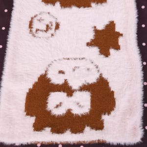 May include: A close-up of a soft, fluffy, off-white textile with brown owl and star designs. The owl has large eyes and a bow tie. The background is a dark brown with pink polka dots. The textile has a plush, fuzzy texture.