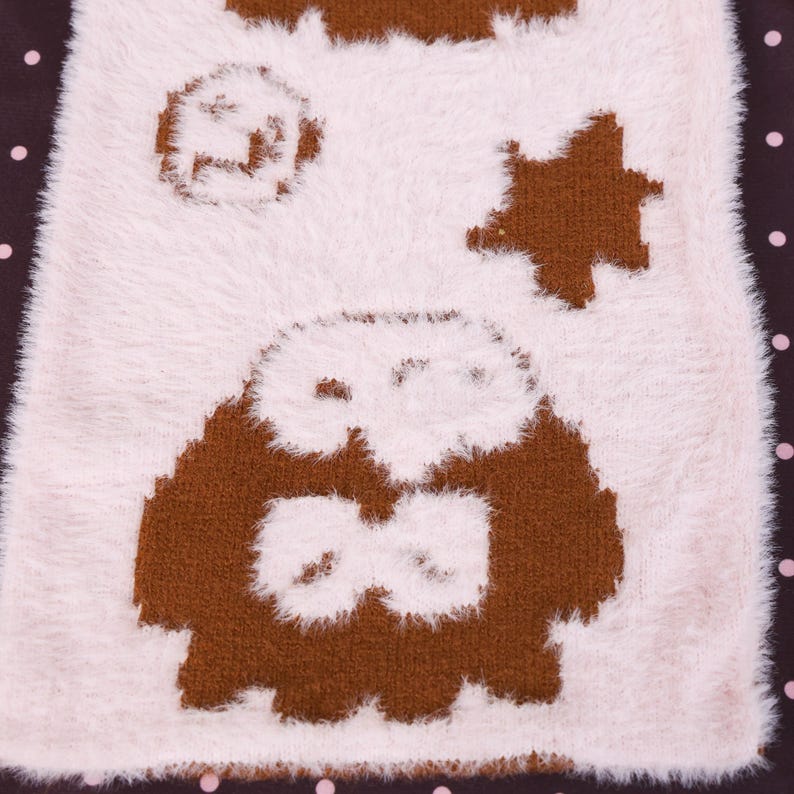 May include: A close-up of a soft, fluffy, off-white textile with brown owl and star designs. The owl has large eyes and a bow tie. The background is a dark brown with pink polka dots. The textile has a plush, fuzzy texture.