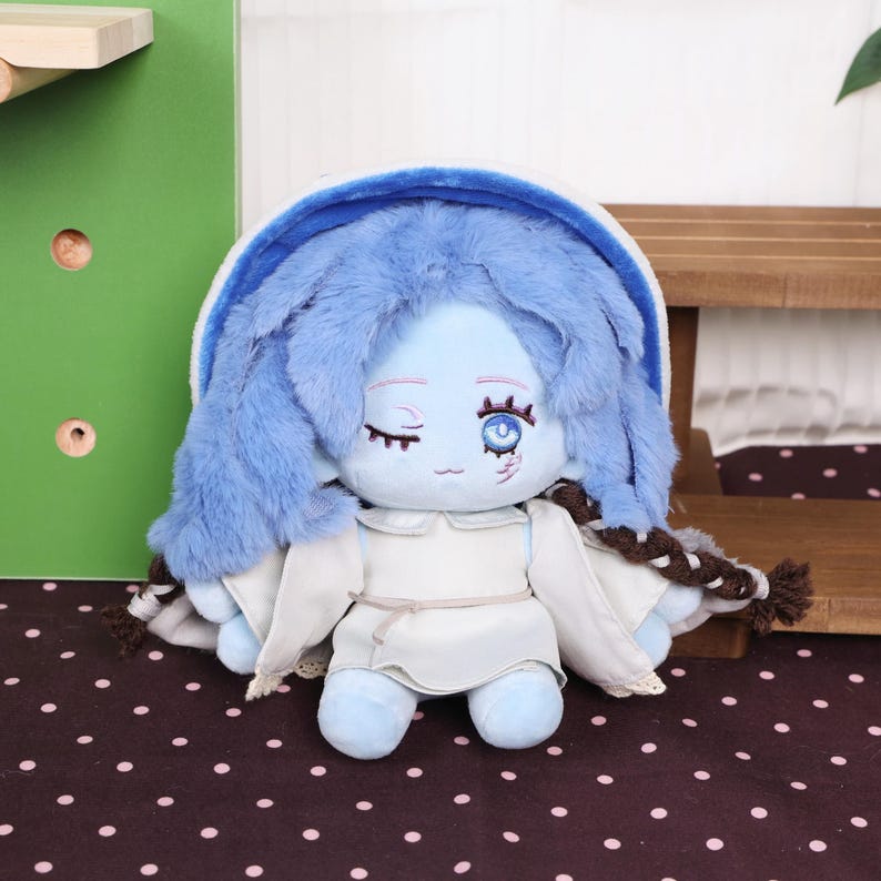 May include: A plush doll with blue skin, blue fluffy hair, and a white dress. The doll has one eye open and one closed, with a small scar detail. The doll is a soft toy.
