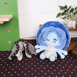 May include: A blue plush doll with blue hair and a white dress. The doll has closed eyes and a serene expression. A matching blue shell-shaped accessory and a gray and brown braided accessory are also present.