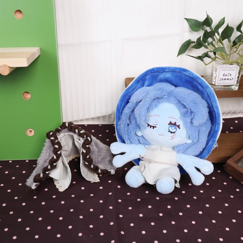 May include: A blue plush doll with blue hair and a white dress. The doll has closed eyes and a serene expression. A matching blue shell-shaped accessory and a gray and brown braided accessory are also present.