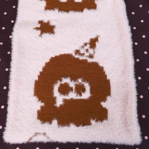 May include: A close-up of a white and brown fuzzy textile with a repeating pattern. The pattern features a brown ghost-like figure with a star and other shapes. The background is a dark brown with white polka dots.